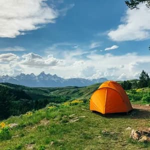 Orange dome tent pitched on grass overlooking rolling green hills, wildflowers, and snow-capped mountains under a blue sky with white clouds.