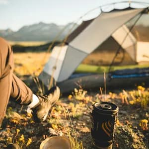 Jet Boil camping stove with black and gold branding on ground next to tent in mountain landscape at sunset